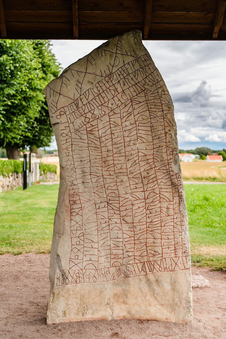 Carved ancient rune stone under wooden shelter with green field.