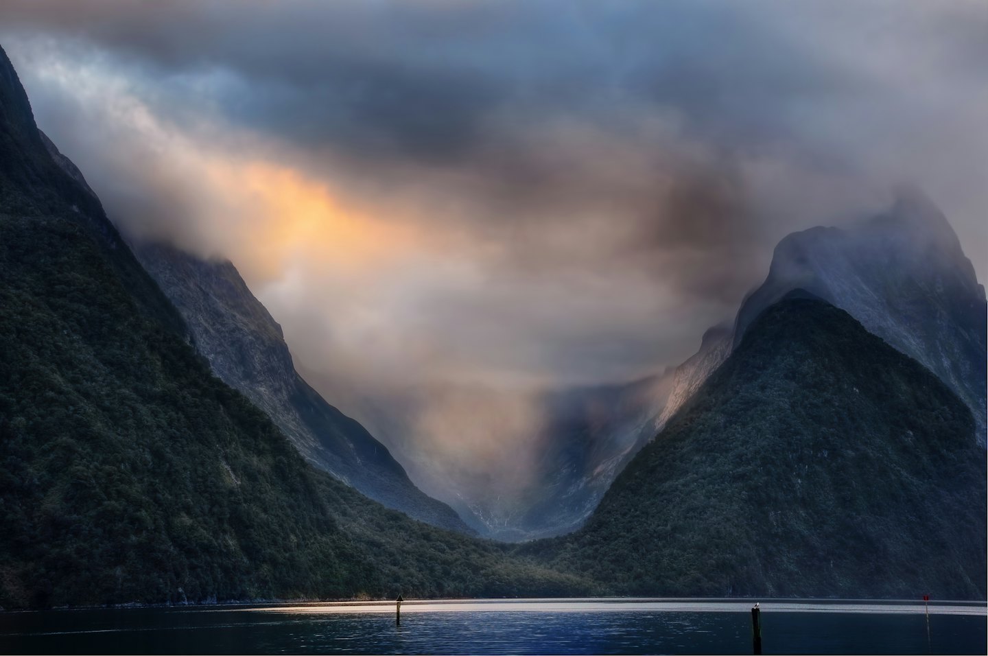 Steep forested mountains rise from a dark fjord under cloudy skies.